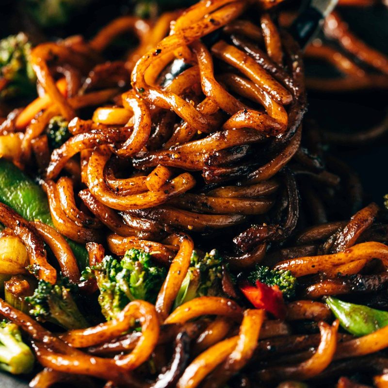 Udon Noodles mixed with fried vegetables and some black pepper in a cooking pan.